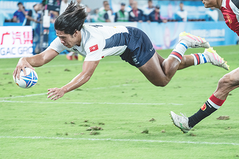 ◎ 十一月十二日,第十五屆全國運動會(十五運會)七人制橄欖球賽事在香港啟德體育園主場館開賽。在首日進行的男子小組循環賽中,香港隊以二十四比十二戰勝山東隊,首戰告捷。圖為香港隊選手房傑鋒(白衣)躍起達陣。(中新社記者侯宇 攝)